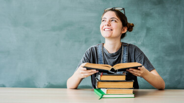 L&auml;chelnde Studentin sitzt an einem Tisch mit einem aufgeschlagenen Buch und einem Stapel B&uuml;cher vor einer gr&uuml;nen Tafel.
