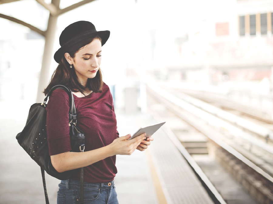 Frau mit Hut steht mit Tablet am Bahnhof.