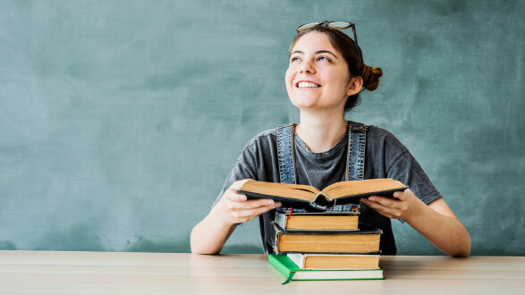 L&auml;chelnde Studentin sitzt an einem Tisch mit einem aufgeschlagenen Buch und einem Stapel B&uuml;cher vor einer gr&uuml;nen Tafel.