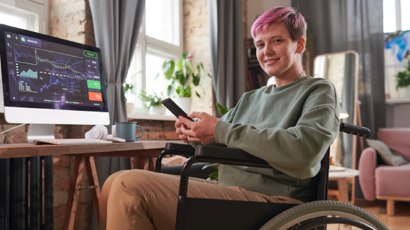 Junge Frau mit kurzen pinken Haaren sitzt mit Handy in der Hand im Rollstuhl vorm Computer, auf dem eine Seite mit Aktienkursen geöffnet ist. Sie lächelt in die Kamera.