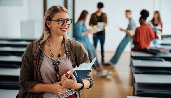 Junge Frau mit Brille steht l&auml;chelnd in einem Klassenraum und h&auml;lt Unterlagen in der Hand. Im Hintergrund unterhalten sich mehrere Studierende in kleinen Gruppen.