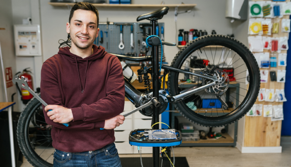 Junger Mann mit Hoodie und Werkzeug in der Hand steht in einer Fahrradwerkstatt und lächelt in die Kamera.