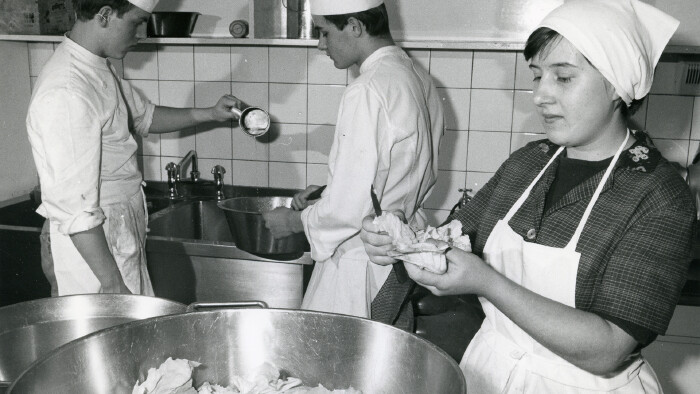 Schwarz-wei&szlig;-Bild aus dem Gastgewerbe in Gastein, Salzburg, 1967: K&ouml;chin mit Sch&uuml;rze und Kopftuch schneidet Blattsalat, im Hintergrund zwei junge K&ouml;che.