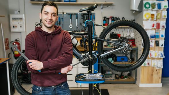 Junger Mann mit Hoodie und Werkzeug in der Hand steht in einer Fahrradwerkstatt und lächelt in die Kamera.