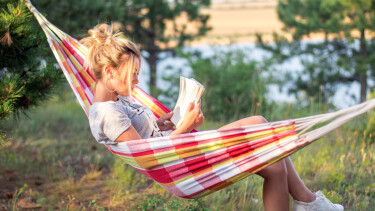 Junge Frau liegt in einer H&auml;ngematte im Gr&uuml;nen am See oder Fluss und liest ein Buch