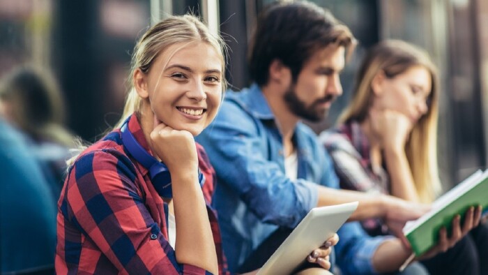 Junge Frau mit Tablet in der Hand l&auml;chelt in die Kamera, im Hintergrund unscharf sitzen ein weiterer Mann und eine Frau mit Unterlagen in der Hand